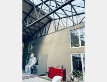 Industriehalle mit transparentem Dach und sichtbarer Stahlkonstruktion. Im Vordergrund eine bemalte Skulptur, ein rotes Sideboard, Zimmerpflanzen sowie eine Wand mit Fenstern und einer Tür.