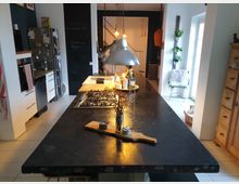 Moderne Wohnküche mit einer zentralen schwarzen Kücheninsel, Gaskochfeld und einer Hängelampe darüber. Im Hintergrund ein großer Kühlschrank, weiße Schränke, eine Tafelwand und eine helle Sitzecke mit Pflanzen an der Wand neben einem Fenster.