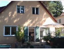 Ein zweistöckiges Einfamilienhaus mit heller Putzfassade und braunem Satteldach. Im Vordergrund befindet sich eine Terrasse mit Pflanzen in Töpfen, einem Tisch mit Stühlen sowie einem Sonnenschirm.