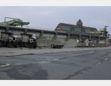 Industriegebiet mit gepflasterter Straße im Vordergrund, mehreren geparkten Lastwagen und einem hohen Gebäude im Hintergrund mit der Aufschrift „BEHALA Westhafen“. Über dem Gelände verläuft eine Stahlkonstruktion mit einem großen grünen Kran.