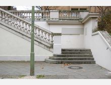 Außenbereich mit einer Treppe aus Beton und einem Geländer mit Balustraden, das zu einer höher gelegenen Plattform führt. Rechts und links der Treppe befinden sich weiße Wände, im Vordergrund ein Gehweg mit Pflastersteinen und vereinzelt liegendem Laub.
