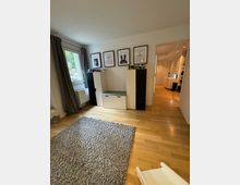 Ein helles Kinderzimmer mit Holzboden, einem grauen Teppich und weißen Möbeln. An der Wand hängen vier gerahmte Bilder mit kindgerechten Motiven, ein großes Fenster mit Vorhängen befindet sich links, und eine Tür führt in einen angrenzenden Flur.