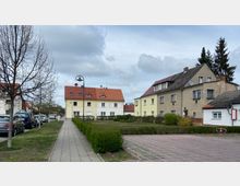 Fußweg in einer Wohnsiedlung mit parkenden Autos auf der linken Seite, mehrstöckige Wohnhäuser mit roten Ziegeldächern im Hintergrund, sowie Hecken und ein gepflegter Vorgarten. Der Himmel ist bewölkt, und Bäume säumen den Weg.