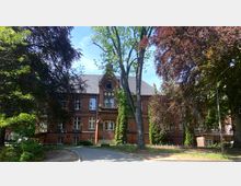 Backsteingebäude im neogotischen Stil, umgeben von großen Bäumen und Grünflächen. Vor dem Gebäude führt ein gepflasterter Weg über einen Hof.