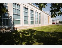 Außenansicht eines weißen Gebäudes mit großen rechteckigen Fenstern, die über mehrere Etagen reichen. Im Vordergrund befindet sich eine grüne Rasenfläche mit Schatten von Bäumen, im Hintergrund sind weitere moderne Gebäude sichtbar.