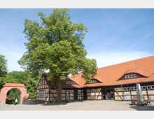 Fachwerkgehöft mit roten Ziegeldächern und hellen Fassaden, umgeben von einem gepflasterten Innenhof; ein großer Baum steht vor dem Hauptgebäude, und links ist ein rotes Backsteinbogenportal zu sehen, das zu einer Grünanlage führt.