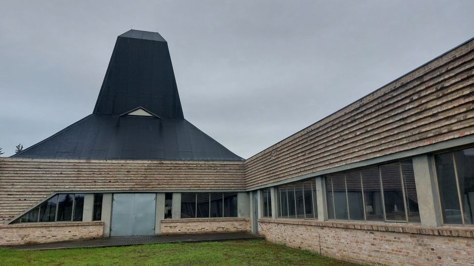 Ein modernes Gebäude mit einer dunklen, pyramidenartigen Dachstruktur und Wänden aus horizontal angeordneten Ziegelsteinen. Große Fensterfronten und ein Eingang aus Metall prägen die Fassade, umgeben von einer Wiese unter einem bedeckten Himmel.