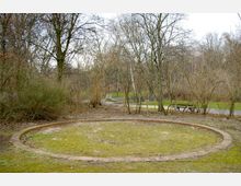 Kreisförmige, mit Backsteinen eingefasste Fläche auf vermoostem Boden in einem Park mit kahlen Bäumen und Sträuchern; im Hintergrund ein Gehweg, eine Bank und vereinzelte grüne Grasflächen.