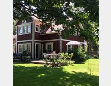 Ein rotes, zweigeschossiges Holzhaus im skandinavischen Stil mit weißen Fensterrahmen und Schrägdach, umgeben von einem Garten mit Rasen, Bäumen und einer Terrasse mit Sitzmöbeln und Sonnenschirm. Im Vordergrund stehen Pflanzen und Sträucher, die den Garten zieren.