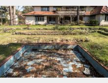 Großer Garten vor einem zweistöckigen Haus mit rot gedecktem Dach, darunter eine Terrasse mit Balustrade. Im Vordergrund ist ein leerer, verwitterter Pool mit roten Ziegeln und blauen Farbresten sichtbar, umgeben von Gras und steinernen Gartenbeeten.