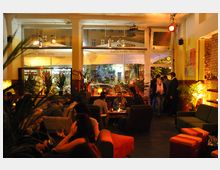 Ein gemütliches Café oder Bar mit warmem, gedimmtem Licht, Sitzmöbeln wie Sofas und Sesseln sowie Pflanzen als Dekoration. Im Hintergrund sind große Fenster und eine lebhafte Atmosphäre mit mehreren Personen sichtbar.