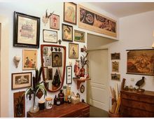Vintage-inspirierter Raum mit dekorativen Wänden, die mit verschiedenen gerahmten Illustrationen, Gemälden und Zeichnungen bedeckt sind. Im Raum befinden sich ein Holzspiegel, ein Sideboard mit Vasen und Keramiken sowie rustikale Dekoelemente wie Tierpräparate und antike Gegenstände.