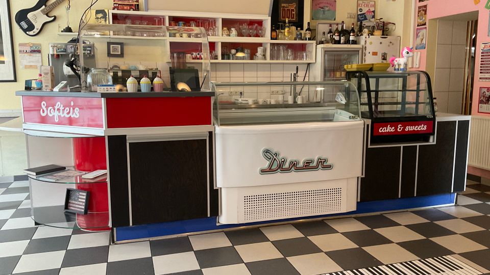 Innenansicht eines Retro-Diners mit schwarz-weißem Schachbrettboden und Verkaufstheke mit Glasvitrinen für Softeis und Kuchen. Hinter der Theke hängen Vintage-Schilder und Regale mit Gläsern und Flaschen; an der Wand ist eine E‑Gitarre befestigt.