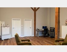 Innenraum eines Büros mit Holzbalken an der Wand, zwei weißen Türen, hellem Laminatboden und neutralen Wänden. Im Raum stehen ein Schreibtisch mit Bürostuhl, zwei grüne Sessel sowie ein Drucker und mehrere Schränke.