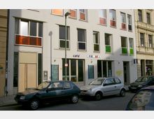 Straßenansicht eines modernen Gebäudes mit hellgrauer Fassade und farbigen Fensterflächen in Orange und Grün. Im Erdgeschoss befindet sich ein Café mit handgeschriebenen Tafeln, daneben parkende Autos auf der Straße.