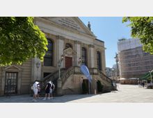 Alt-Text: Historisches Gebäude mit klassizistischer Architektur, einer breiten Freitreppe und mehreren Bogenfenstern, darüber eine Inschrift. Im Vordergrund stehen Menschen unter Bäumen, im Hintergrund ein Gerüst an einem benachbarten Gebäude.