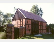 Fachwerkscheune mit rotem Ziegeldach und Wänden aus roten Ziegelsteinen und hölzernen Balken, umgeben von einer Ziegelmauer mit Metalltoren; im Hintergrund Bäume und eine grüne Wiese.