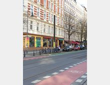 Straßenszene mit einer Altbau-Fassade mit roten und weißen Elementen, im Erdgeschoss befinden sich Geschäfte mit Schaufenstern. Davor stehen Fahrräder an einem Fahrradständer, parkende Autos und eine Straße mit Straßenbahnschienen sowie einem markierten Fahrradweg.