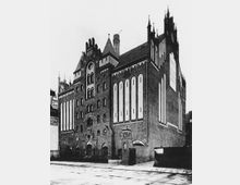 Historisches Backsteingebäude mit neugotischen Architekturelementen, darunter spitze Türme, Spitzbögen und dekorative Fensteröffnungen; das Gebäude hat mehrere hohe, schmale Fensterfassaden und befindet sich in einer städtischen Umgebung.