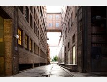 Schmaler, gepflasterter Innenhof zwischen zwei parallelen Backsteingebäuden mit großen, rechteckigen Fenstern; im Hintergrund verbindet ein oberer Steg die Gebäude, und am Ende des Hofes ist Tageslicht sowie ein Baum sichtbar.