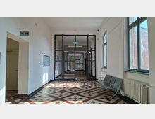 Heller Innenflur in einem öffentlichen Gebäude mit weiß gestrichenen Wänden, großen Fenstern rechts und einer Sitzbank neben einem Heizkörper. Der Boden ist mit rot-weiß gemusterten Fliesen ausgelegt, und am Ende des Gangs führen dunkle Glasrahmentüren in weitere Korridore; links weist ein WC-Schild auf einen Nebenraum.