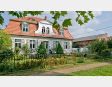 Ein zweistöckiges Haus mit weißer Fassade und roten Dachziegeln, umgeben von einem Garten mit Sträuchern und Bäumen. Vor dem Haus steht ein schmiedeeiserner Zaun, und im Hintergrund ist ein rotes Backsteingebäude zu sehen.