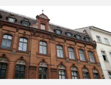 Historisches Backsteingebäude mit roten und gelben Ziegeln, verzierten Rundbogenfenstern und kunstvollen Reliefs oberhalb der Fenster; das Dach ist mit dunkelbraunen Dachschindeln gedeckt und verfügt über mehrere kleine Dachgauben.
