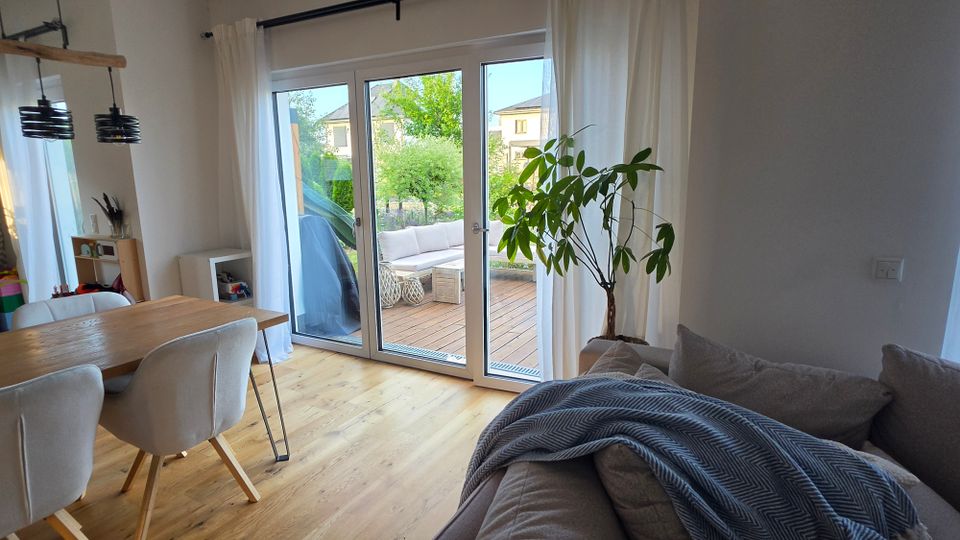 Heller Wohn- und Essbereich mit Holzboden, Esstisch mit Polsterstühlen, Sofa und großer Zimmerpflanze. Eine breite Glas-Schiebetür mit weißen Vorhängen führt auf eine Holzterrasse mit Sitzmöbeln und Blick in einen grünen Garten.