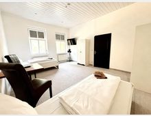 Helles, schlichtes Gästezimmer mit zwei Einzelbetten, grauem Teppichboden und weißer Holzdecke. Zwei Fenster mit Rollläden, ein kleiner Tisch mit Stuhl, ein Schrank sowie ein wandmontierter Fernseher und eine dunkle Tür sind zu sehen.