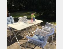 Weißer Gartentisch mit vier weißen Korbstühlen, die blaue Kissen haben, auf einer gepflasterten Terrasse in einem Garten. Auf dem Tisch stehen eine Vase mit rosa Tulpen, eine Glasflasche mit Getränk und mehrere Gläser.