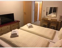 Ein einfach eingerichtetes Hotelzimmer mit zwei Einzelbetten, auf denen gefaltete Handtücher liegen. Im Raum befinden sich eine Kommode mit Fernseher, ein Schreibtisch mit zwei Holzstühlen und ein Spiegel, sowie eine Tür zum angrenzenden Bereich.