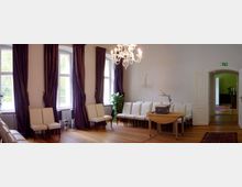 Ein heller, eleganter Raum mit Holzfußboden, weißen Wänden und hohen Fenstern, die mit dunklen Vorhängen dekoriert sind. Der Raum ist mit weißen Stühlen, einem kleinen Holztisch, einem Kronleuchter und einer offenen Tür, die in einen angrenzenden Raum führt, ausgestattet.