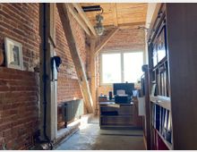 Ein Büro in einem rustikalen Raum mit freiliegenden Ziegelwänden, Holzbalken und einer Holzdecke. Im Vordergrund stehen ein Regal mit Aktenordnern und ein Schreibtisch mit Computer sowie Drucker nahe einem Fenster, das Tageslicht hereinlässt.