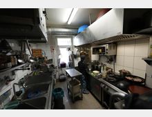 Schmale Großküche mit weißen Fliesenwänden, Edelstahl-Arbeitsflächen und großem Dunstabzug über einem Gasherd mit mehreren Pfannen und Töpfen. Links sind Spüle und Regale mit hängenden Küchenutensilien zu sehen; am Ende des Raums befindet sich ein Fenster und eine Lüftungsleitung unter der Decke.