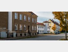 Straßenszene mit mehrstöckigen Wohngebäuden, darunter ein braunes Altbauhaus und ein weiß verputztes Gebäude mit roten Ziegeldächern. Im Vordergrund sind herbstlich gefärbte Bäume, parkende Autos und ein Gehweg zu sehen.