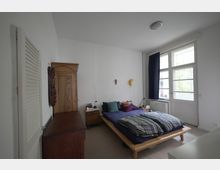 Schlafzimmer mit einem niedrigen Holzbett, das blaues Bettzeug und bunte Kissen hat. An der rechten Wand befinden sich große Fenster mit blickdichten, blauen Vorhängen, daneben ein Heizkörper. Links steht ein Holzschrank und eine Kommode, die Wände sind weiß gestrichen.