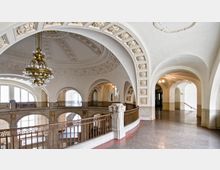 Heller Innenraum eines historischen Gebäudes mit dekorativ verzierten Rundbögen, einer reich verzierten Kuppeldecke und einer prunkvollen, goldfarbenen Hängelampe. Ein umlaufendes Geländer mit geometrischem Muster grenzt eine offene Galerieebene ab.