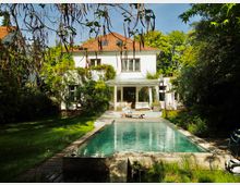 Zweistöckiges weißes Wohnhaus mit roten Dachziegeln, umgeben von einer üppigen Gartenlandschaft. Im Vordergrund befindet sich ein rechteckiger Swimmingpool mit angrenzender Terrasse und Gartenmöbeln.