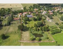 Luftaufnahme einer ländlichen Wohnanlage mit großzügigem Grundstück, umgeben von Feldern und weiteren Häusern. Das Gelände ist von einer roten Linie umrandet, umfasst Grünflächen, Bäume, einen Teich sowie mehrere Gebäude mit roten Dächern, einschließlich einer Photovoltaikanlage.