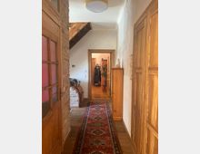 Ein Flur in einem Wohnhaus mit Holzboden und hell verputzten Wänden. Auffällig sind eine gemusterte rote Teppichläufer, Holztüren, eine Ziegelwand sowie eine Holztreppe, die seitlich nach oben führt.