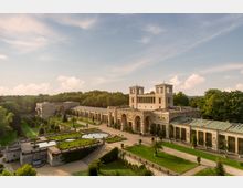 Klassizistisches Schlossgebäude mit Türmen und Arkaden, eingebettet in eine weitläufige Gartenlandschaft mit symmetrischen Brunnenanlagen, Rasenflächen und Baumbestand. Im Hintergrund dichte Wälder unter einem leicht bewölkten Himmel.