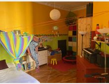 Ein Kinderzimmer mit bunten Wänden in Gelb- und Grüntönen, dekoriert mit einem farbenfrohen Stofftuch, das über einer Ecke des Raums gespannt ist. Es gibt Regale mit Spielzeug, eine Kommode mit einer Stereoanlage, einen kleinen Spielbereich mit Trampolin und Hockern sowie farblich sortierte Würfelregale.