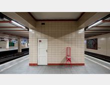 Eine U-Bahn-Station mit cremefarbenen gefliesten Wänden und roten Details. Im Vordergrund eine Wand mit einer weißen Tür und einem roten Gestell, dahinter befinden sich zwei Gleise und ein Bahnsteig mit Informationsschildern und Kunstwerken.