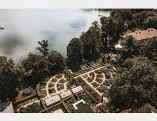 Luftaufnahme einer Landschaft mit einem Garten, der in geometrische Beete unterteilt ist, umgeben von dichtem Grün und einem See im Hintergrund. Rechts befinden sich ein Gebäude mit rotem Dach und weiße Stühle, die in Reihen für eine Veranstaltung aufgestellt sind.