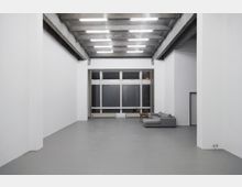 Minimalistischer, leerer Innenraum mit hohen weißen Wänden, einem grauen Betonboden und einer Decke mit sichtbaren Betonträgern und länglichen Deckenleuchten. Am Ende des Raumes befindet sich eine große Fensterfront sowie eine graue Sofaecke mit kleinem Tisch.