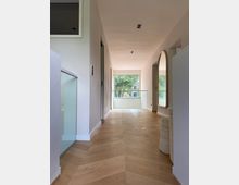 Ein moderner, heller Flur mit Holzfußboden in Fischgrätmuster, weißen Wänden und großen Fenstern am Ende, die einen Blick auf Bäume im Außenbereich ermöglichen. Rechts ist ein großer, ovaler Spiegel montiert, links ist ein Glasgeländer sichtbar.