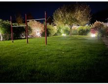 Grünfläche eines Gartens bei Nacht, beleuchtet durch mehrere Bodenlichter. Im Vordergrund steht eine rot-weiße Turnstange, im Hintergrund sind Büsche, Ziergräser und ein einzelner Baum zu sehen.