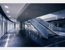 Moderner Innenraum mit einer Metalltreppe und einer geschwungenen Betonwand. Der Boden ist dunkel glänzend, die Beleuchtung erfolgt durch eingelassene Deckenlichter, und an der rechten Seite befinden sich schmale vertikale Fenster.