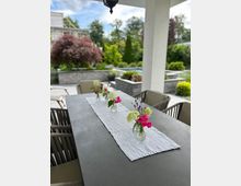 Überdachte Terrasse mit einem langen Esstisch und gepolsterten Gartenstühlen; auf einem weißen Tischläufer stehen mehrere kleine Glasvasen mit Blumen. Im Hintergrund liegt ein modern gestalteter Garten mit niedrigen Steinmauern, Beeten und einem Pool.