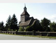 Historische Fachwerkkirche mit steilem Ziegeldach und einem Kirchturm aus Fachwerk und Holz, umgeben von einer niedrigen Ziegelmauer, Bäumen und Grünflächen. Im Vordergrund verläuft eine asphaltierte Straße.